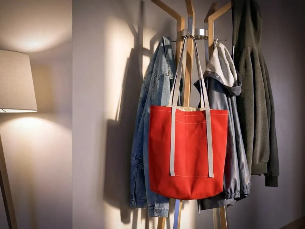 Roter Shopper aus Markisenstoff, hängend an einer Garderobe mit Jacken und warmem Licht im Hintergrund. Stiftung 50 Plus.
