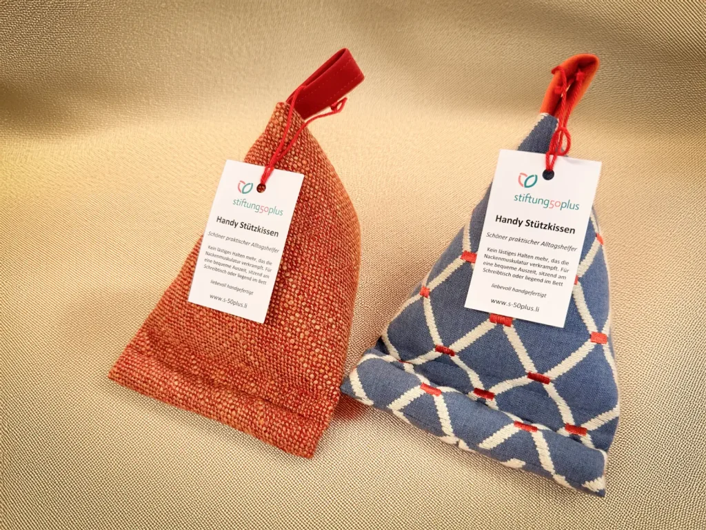 Zwei handwerklich gefertigte Handy-Stützkissen in unterschiedlichen Stoffdesigns, seitlich nebeneinander arrangiert. Stiftung 50 Plus.